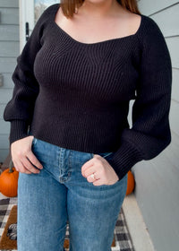 Person wearing a black sweater and blue jeans on a porch with pumpkins.