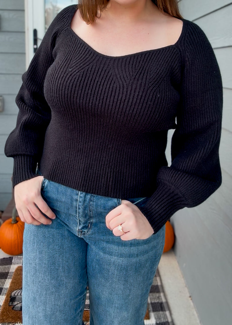 Person wearing a black sweater and blue jeans on a porch with pumpkins.