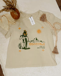 Beige t-shirt with cowboy graphic and 'Cowgirls Do It Better' text on a textured surface.