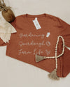 Brown t-shirt with 'Gardening Sourdough Farm Life' text and tag, next to a beaded necklace on a light background.