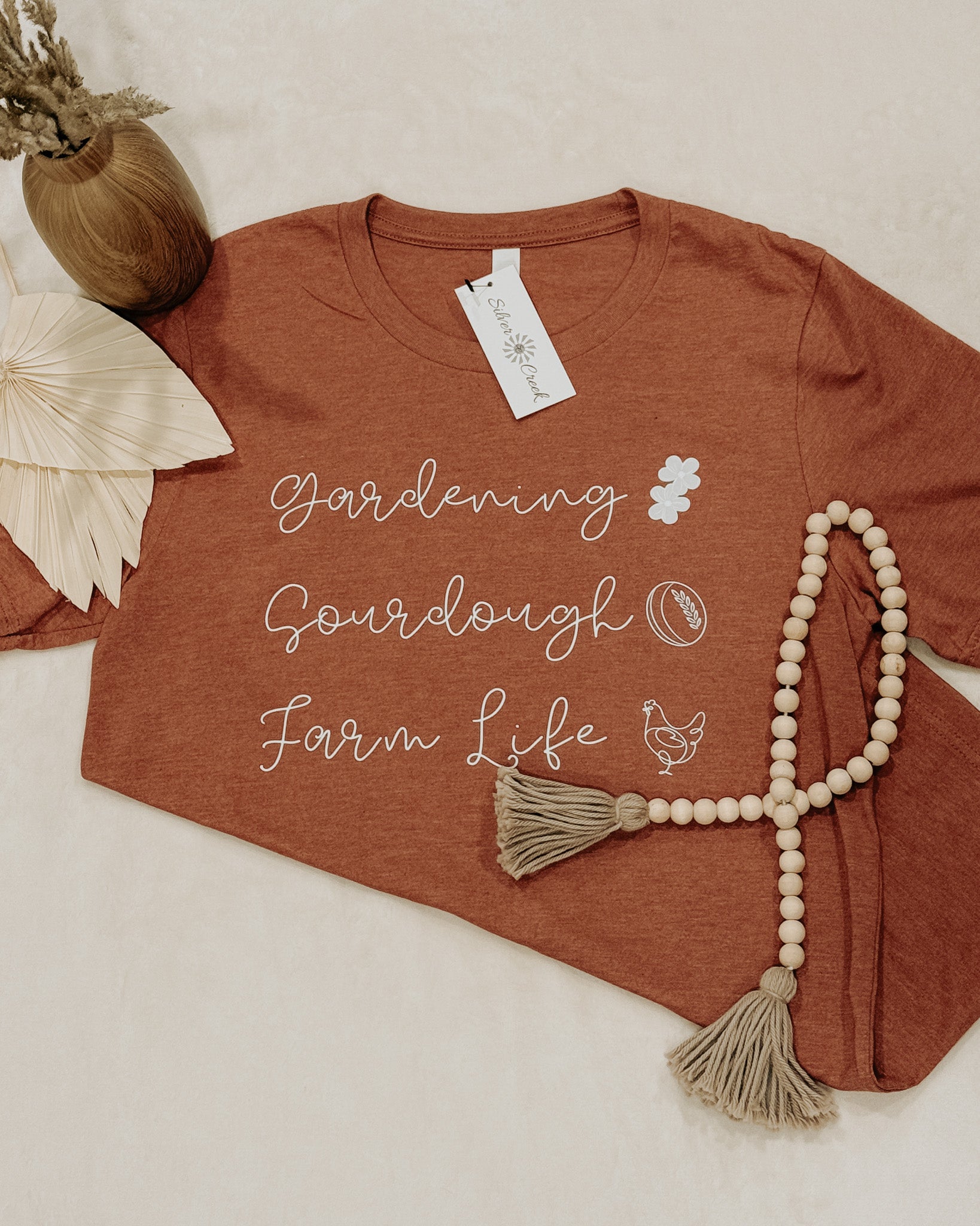Brown t-shirt with 'Gardening Sourdough Farm Life' text and tag, next to a beaded necklace on a light background.